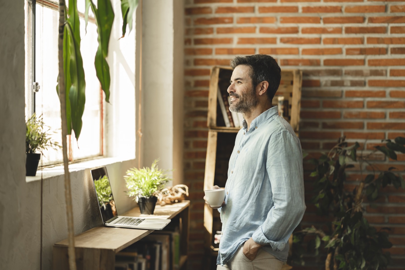 Man at home with coffee, looking out a sunlit window